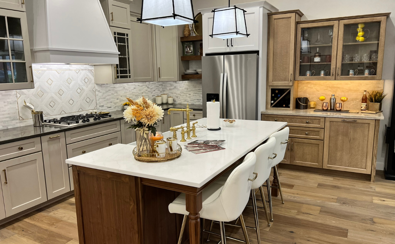 Modern kitchen with a central island, gold faucet, floral decor, light gray cabinets, stainless steel fridge, bar area with wine rack, pendant lights, wood flooring, and warm gold accents.